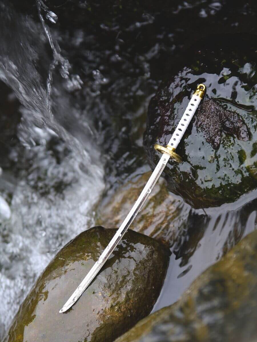 Ichimonji miniature katana, unsheathed and wet, lying on rocks with water droplets, amidst flowing water and a small waterfall.