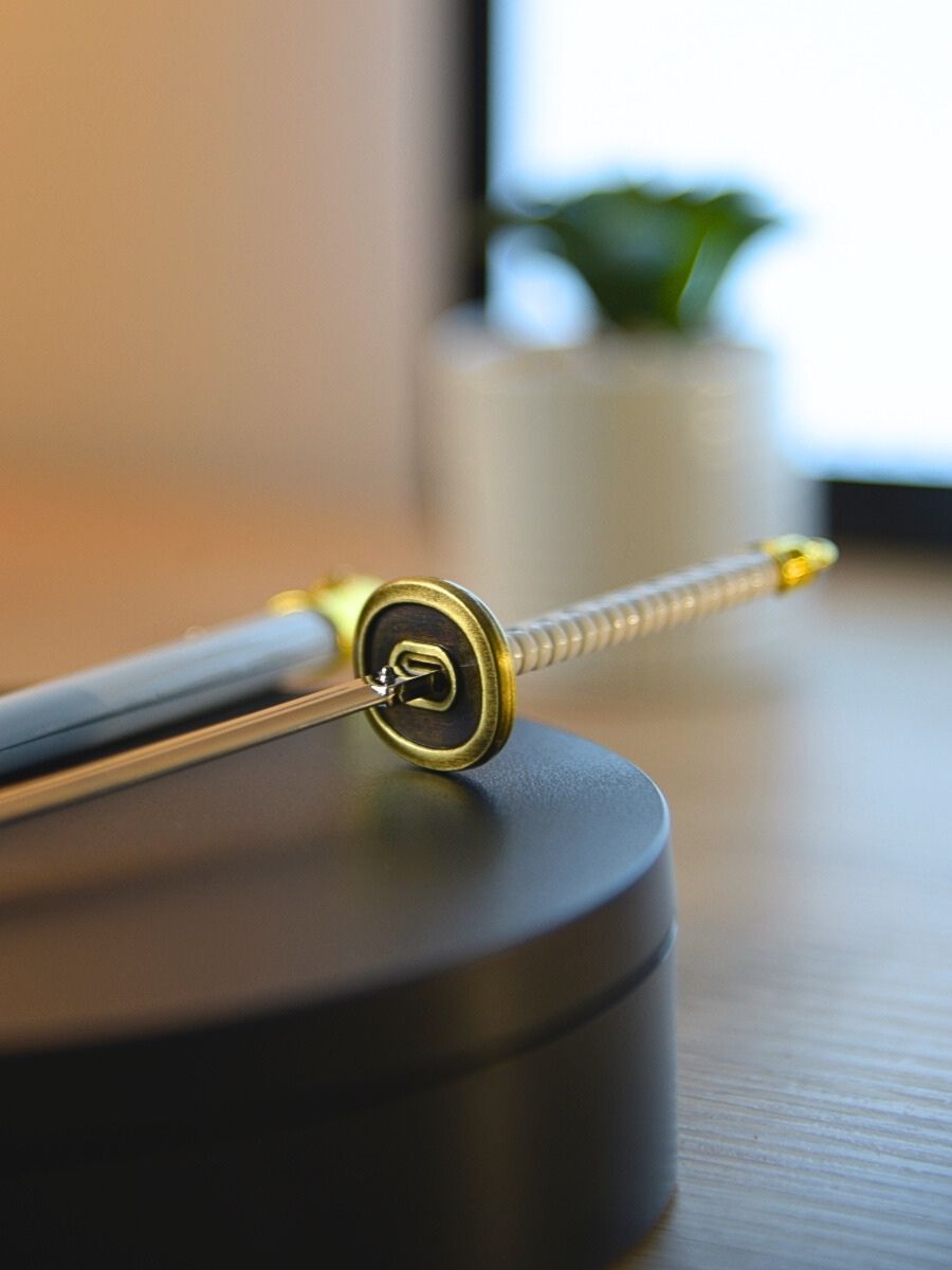Ichimonji miniature katana guard detail on a black display stand, with a blurred background featuring a small cactus indoors.