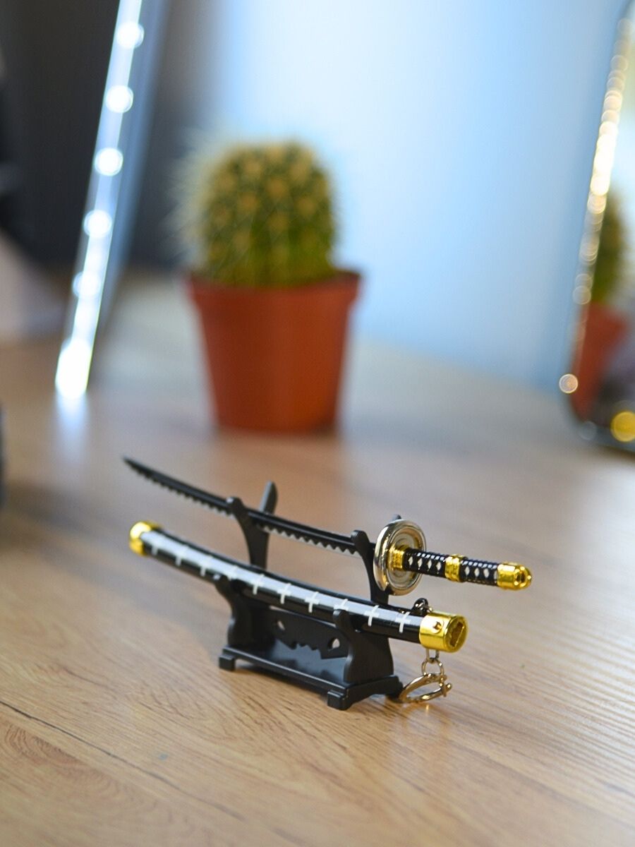 Trafalgar miniature katana from One Piece, displayed without its scabbard, stands gracefully on a desk stand. A delicate touch of ambiance is provided by a small cactus in the background, enhanced by pleasing lighting.