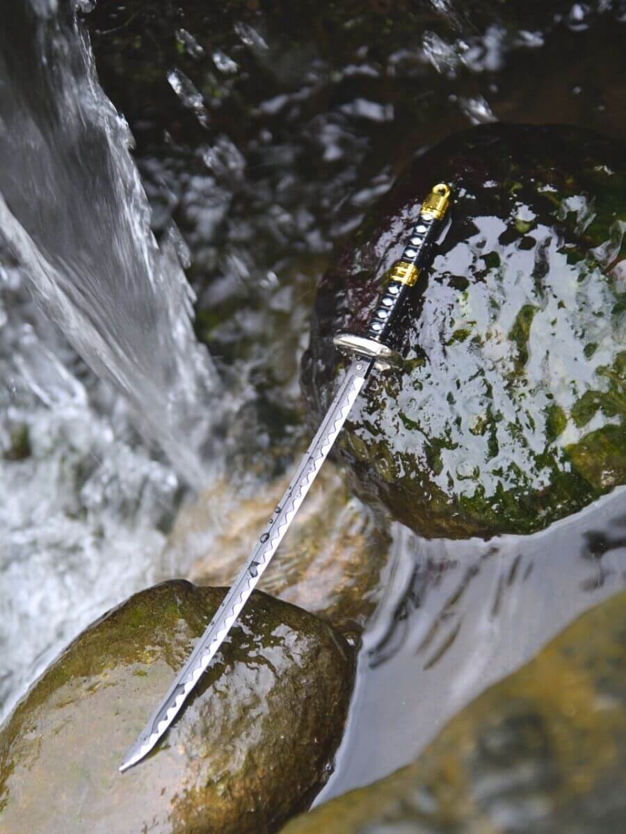 Trafalgar miniature katana from One Piece, without its scabbard, rests on moisture-kissed rocks, creating a riverside ambiance with hints of a nearby waterfall.