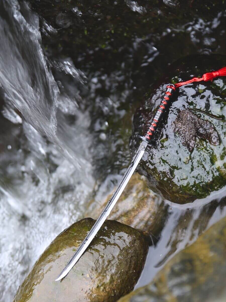 Tanjiro miniature katana, without its scabbard, laid on wet rocks. The scene suggests a riverside ambiance with traces of a gentle waterfall nearby.