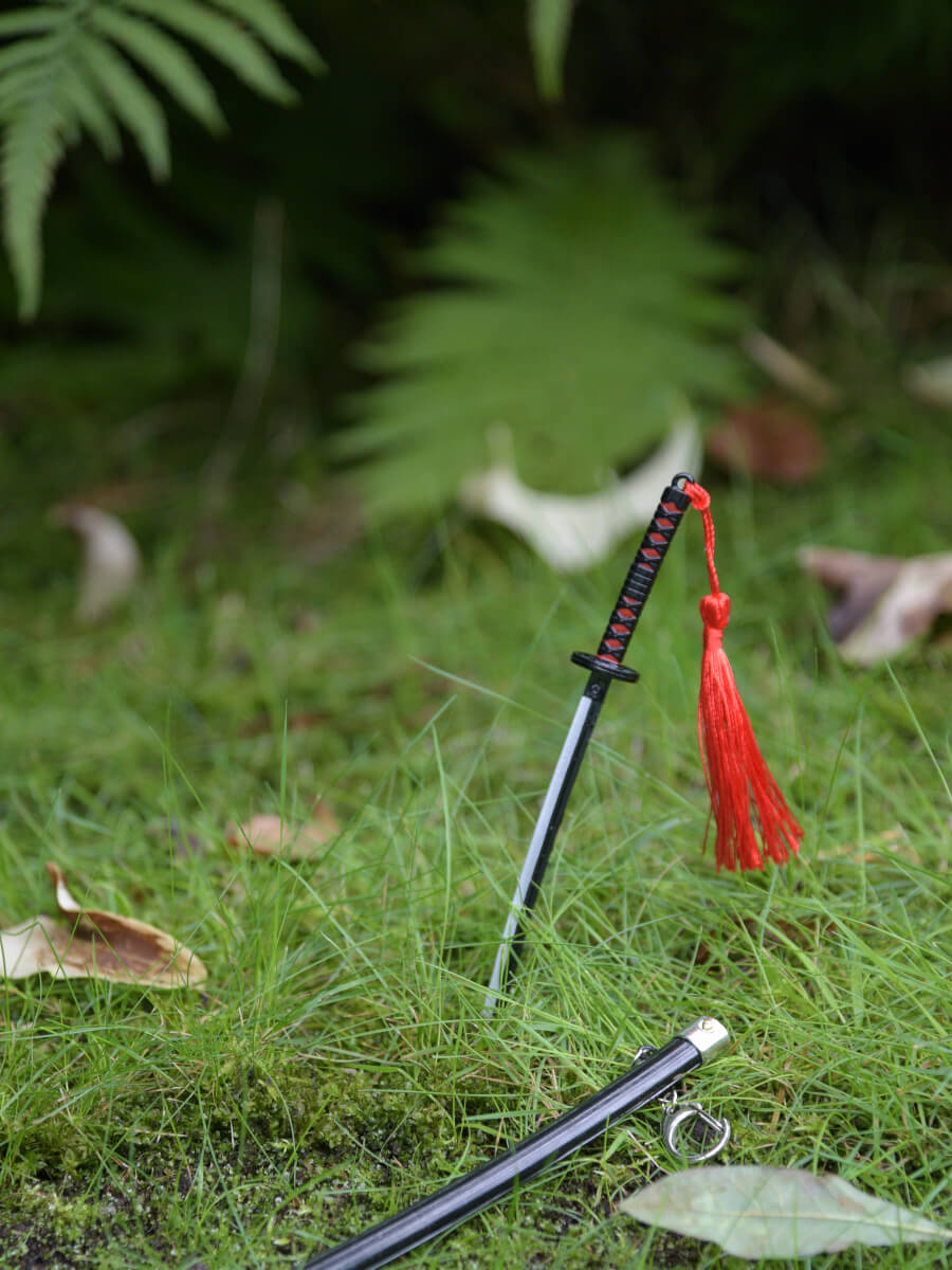 Tanjiro miniature katana from Demon Slayer thrust into the grass, with its scabbard nearby, suggesting a swift unsheathing.